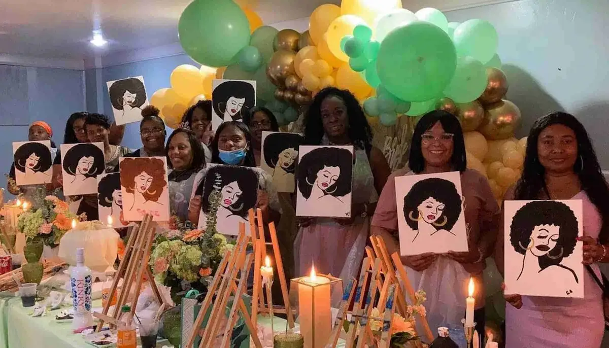 Group of women holding paintings of a woman with Afro hairstyle at a colorful balloon-decorated event.