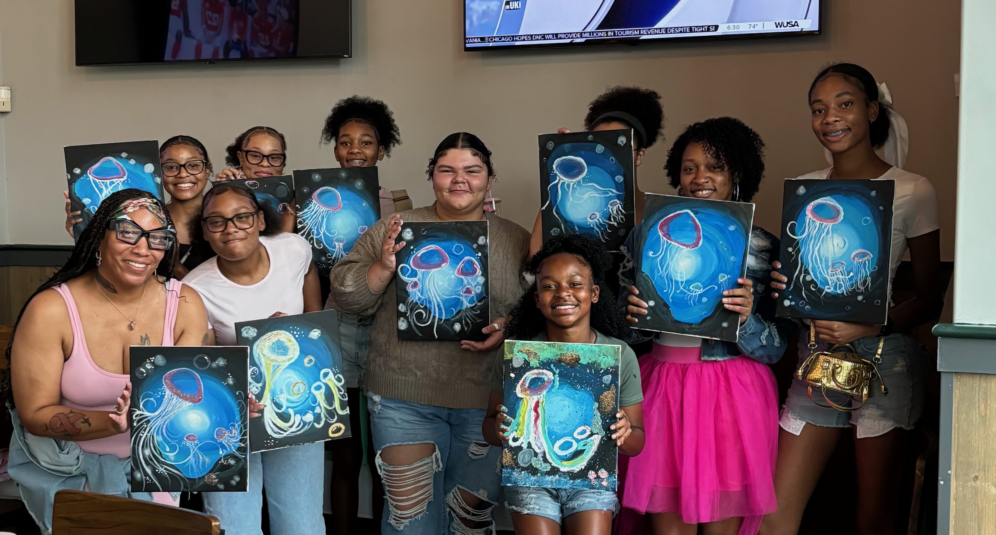 A group of eleven girls holding up paintings of jellyfish in blue underwater scenes, posing indoors.