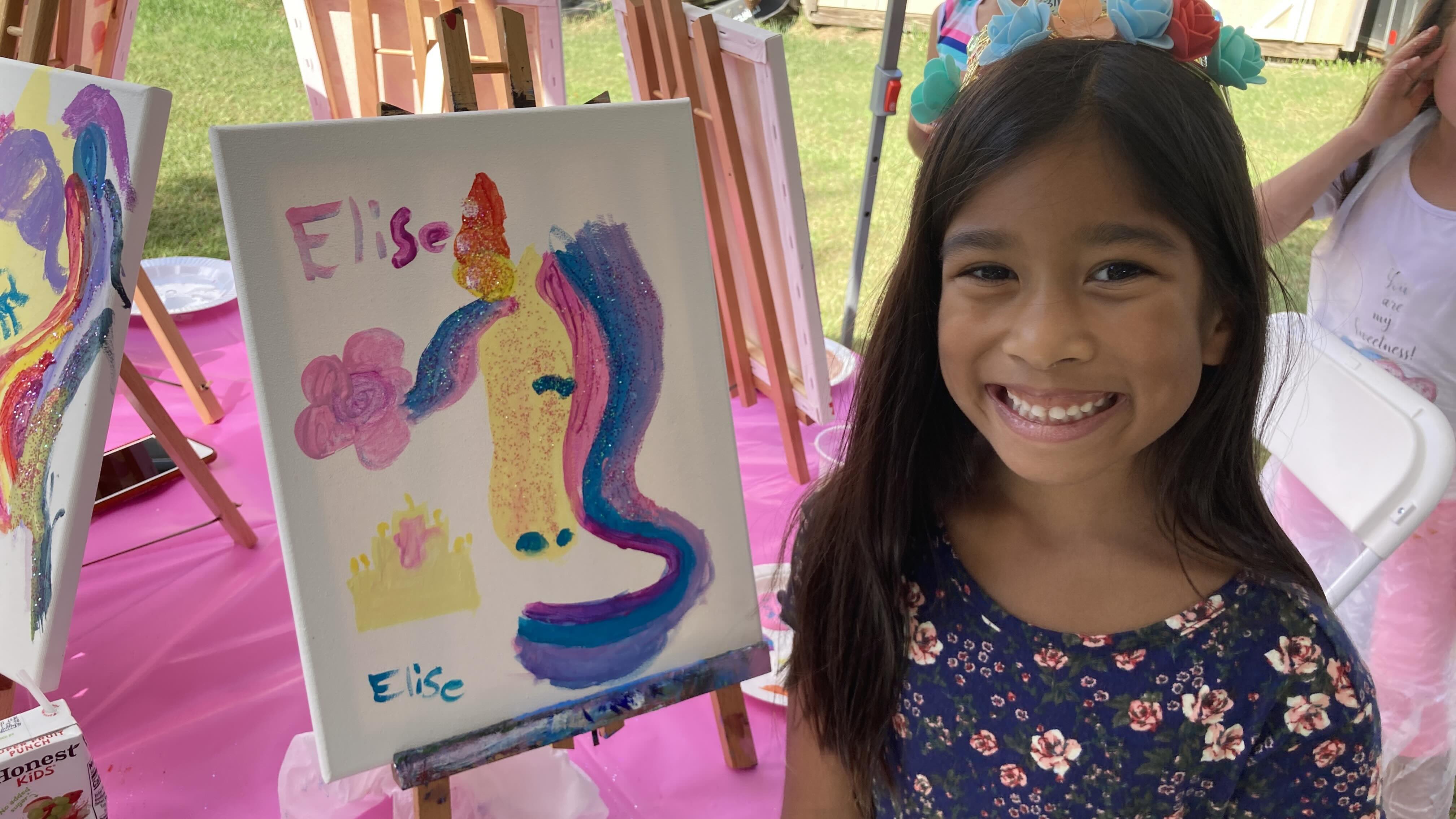 Group of children standing behind a table holding paintings of waterfalls with pink flowers, under a 'Happy Birthday' banner.