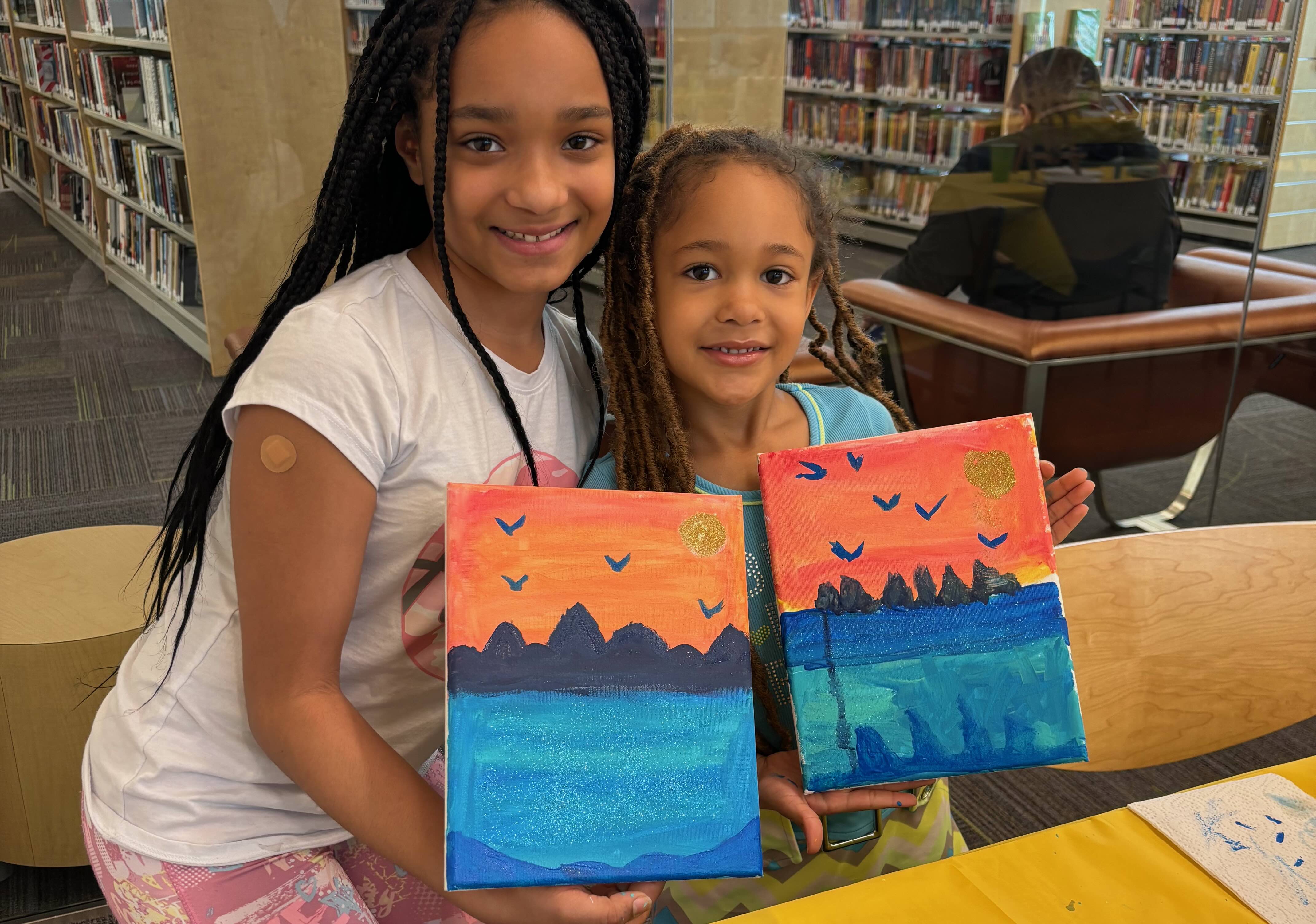 Two smiling children in a library hold paintings of blue water, dark mountains, orange sky, and flying birds, with a glittery sun in the corner.