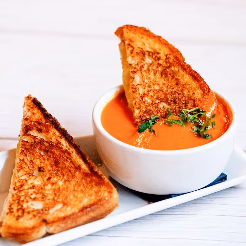 Grilled cheese sandwich halves served with a cup of creamy tomato soup garnished with parsley on a white rectangular plate.