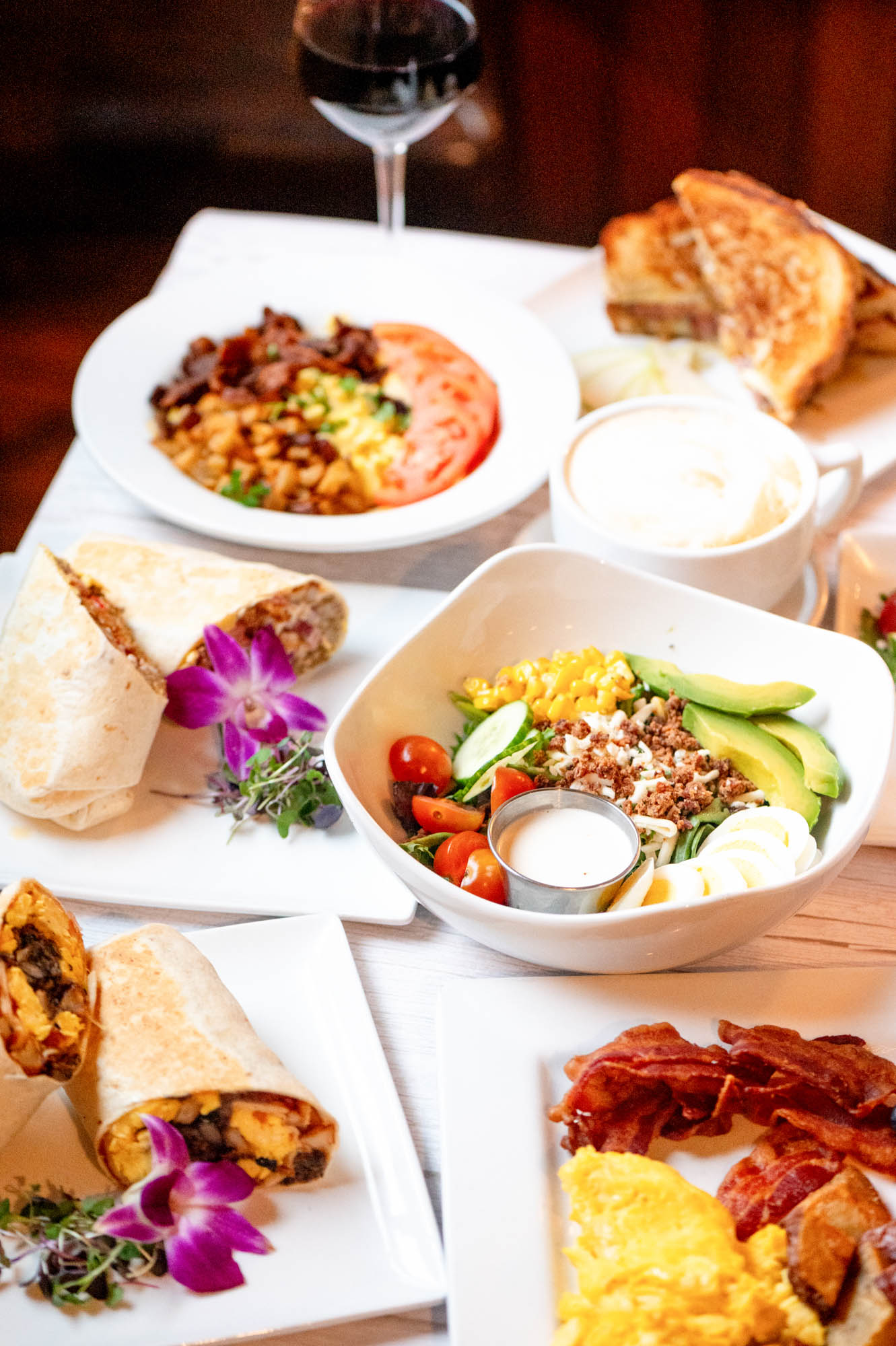 A breakfast spread featuring scrambled eggs with bacon, a salad with avocado, cherry tomatoes, corn, boiled egg slices, and dressing, breakfast burritos garnished with purple flowers, a bowl with scrambled eggs, tomato slices, and potatoes, toasted bread, a cup of coffee, and a glass of red wine.