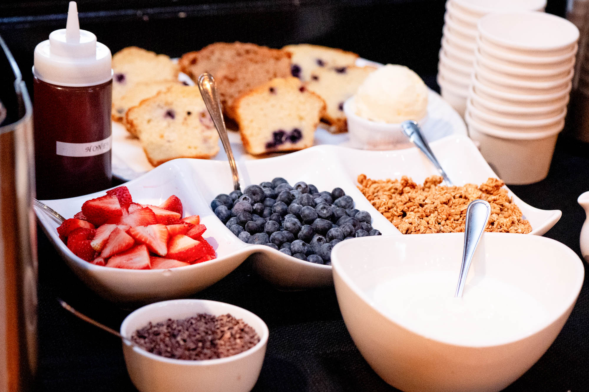 Assorted breakfast toppings including sliced strawberries, blueberries, granola, yogurt, honey, and slices of blueberry muffin.
