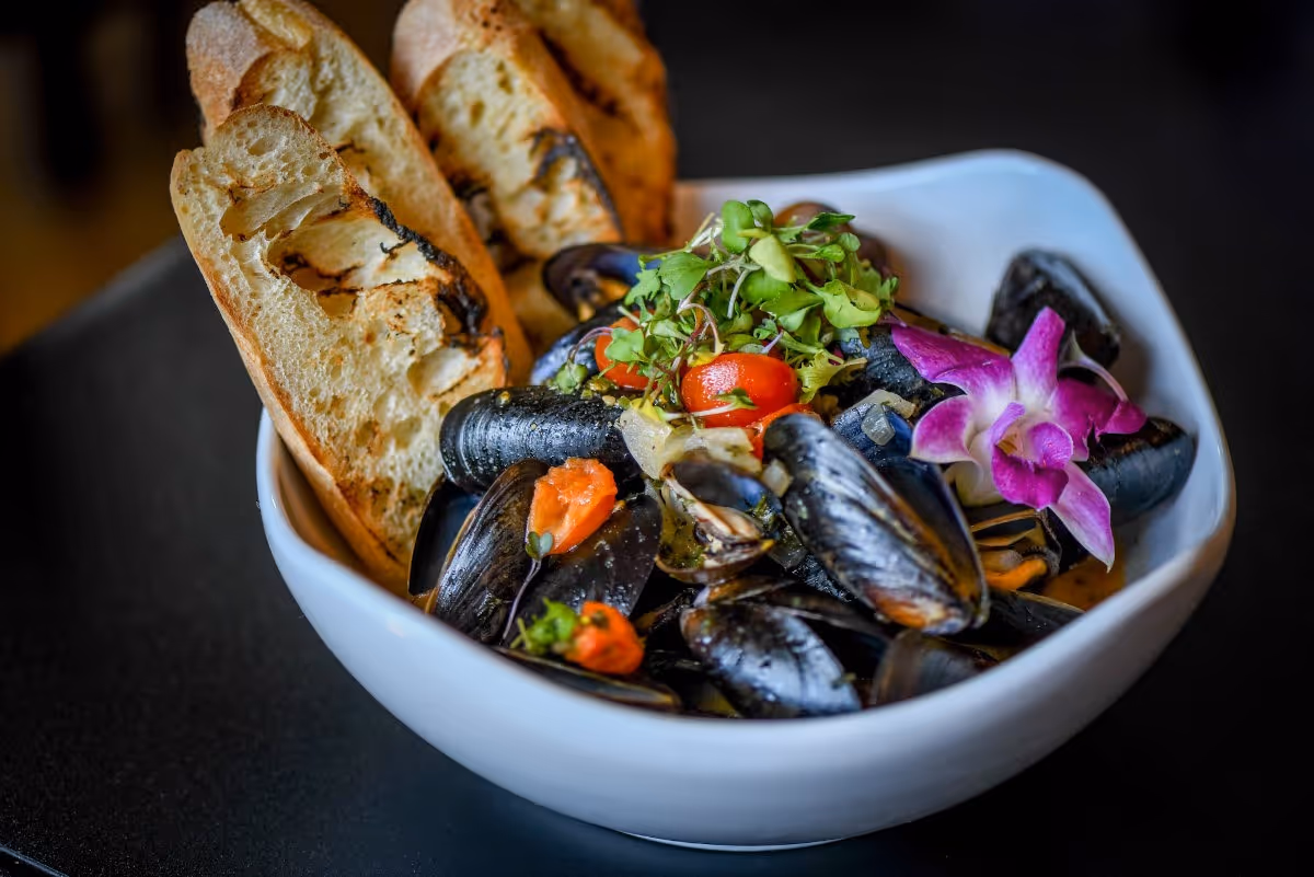 Bowl of cooked mussels garnished with cherry tomatoes, greens, an edible purple orchid, and three slices of grilled bread.