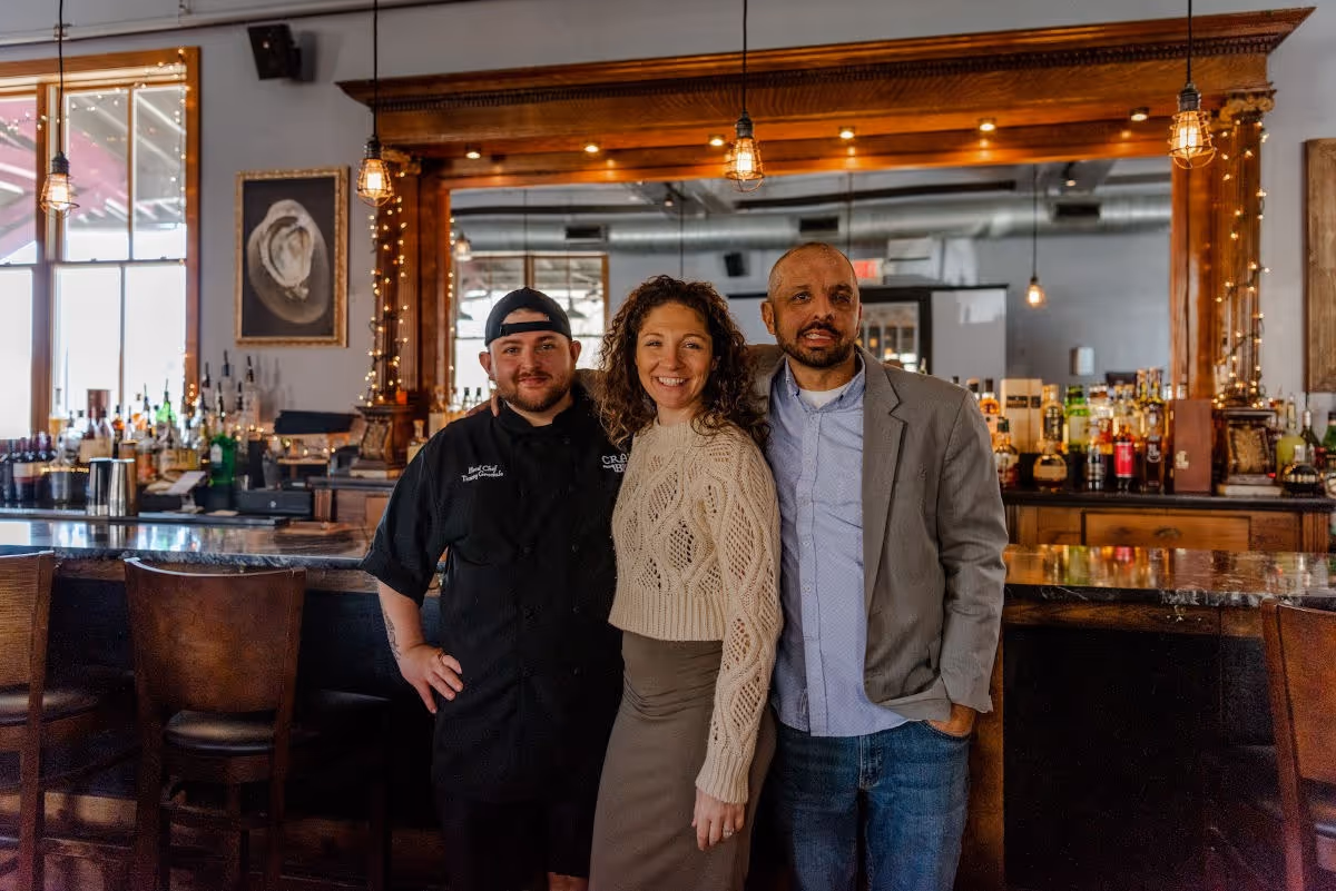Three people standing together and smiling inside a bar with wooden furniture and hanging lights.