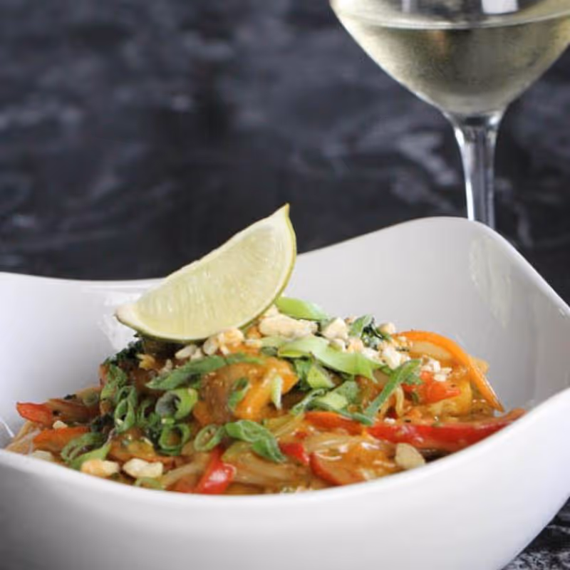 White bowl with Pad Thai garnished with lime wedge, crushed peanuts, and green onions, with a glass of white wine in the background.
