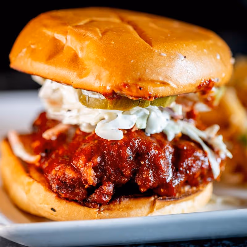 Close-up of a crispy fried chicken sandwich with barbecue sauce, coleslaw, and pickles in a toasted bun.