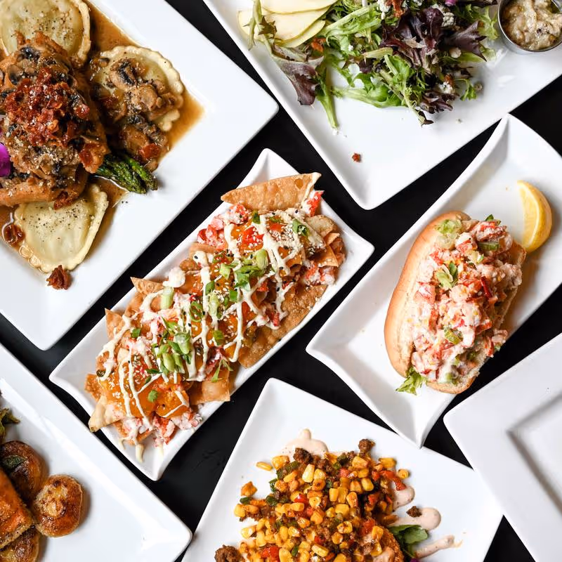 Assorted dishes including ravioli with mushroom sauce, salad with pear slices and dressing, lobster roll with lemon wedge, nachos with toppings, and a corn and vegetable side dish on white plates.