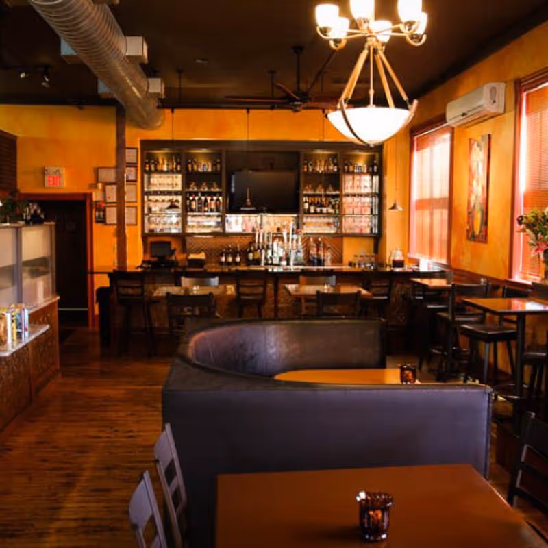 Cozy bar and lounge area with wooden floors, yellow walls, a curved leather booth, tables, and shelves stocked with bottles behind the bar.