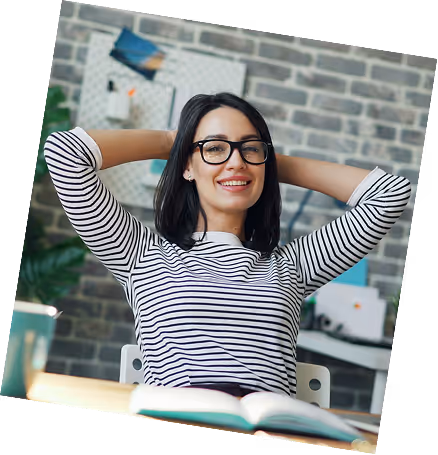 Smiling woman with glasses and striped shirt leaning back with hands behind her head at a desk with an open book.
