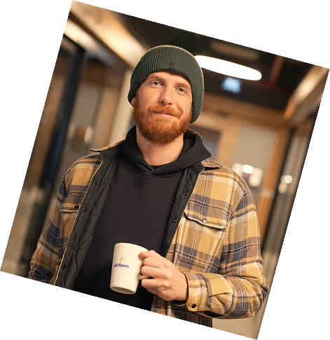 Smiling man with a green beanie and plaid jacket holding a white coffee mug indoors.