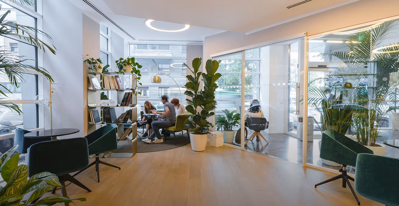 Modern office lounge with green chairs, potted plants, a bookshelf, and people working at a round table near large windows.