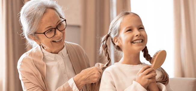 Smiling elderly woman braiding the hair of a happy young girl holding a hairbrush.
