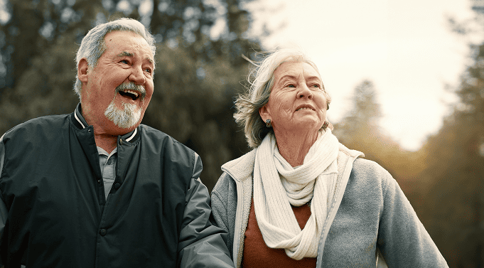 Smiling elderly man and woman outdoors, dressed warmly with trees and sunlight in the background.