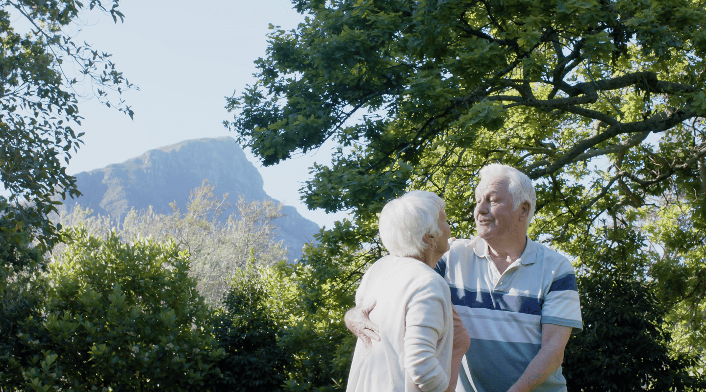 Elderly couple embracing and smiling outdoors with trees and a mountain in the background.