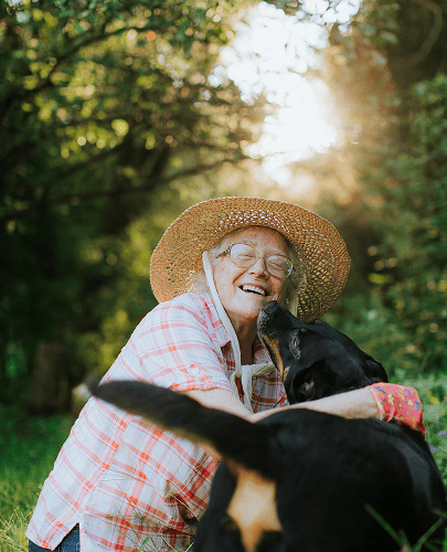 Smiling elderly woman wearing glasses and a straw hat cuddling a black dog outdoors in a sunlit green area.