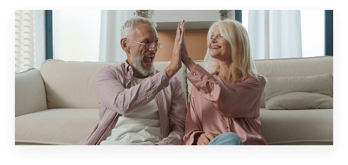Smiling elderly couple with no medicare coverage, sitting on the floor in front of a sofa giving each other a high five.