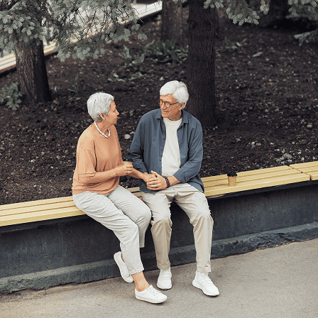 Elderly couple sitting on a wooden bench outdoors, holding hands and smiling at each other.