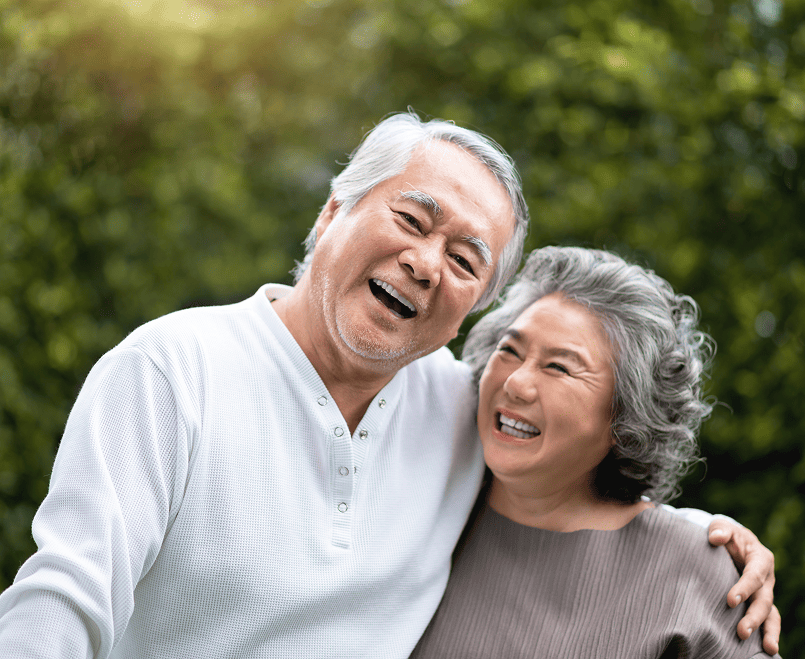 Smiling elderly Asian couple embracing outdoors with greenery in the background.