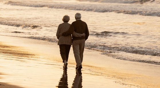 Older couple on medicaid walking arm in arm along the beach at sunset with gentle waves in the background.
