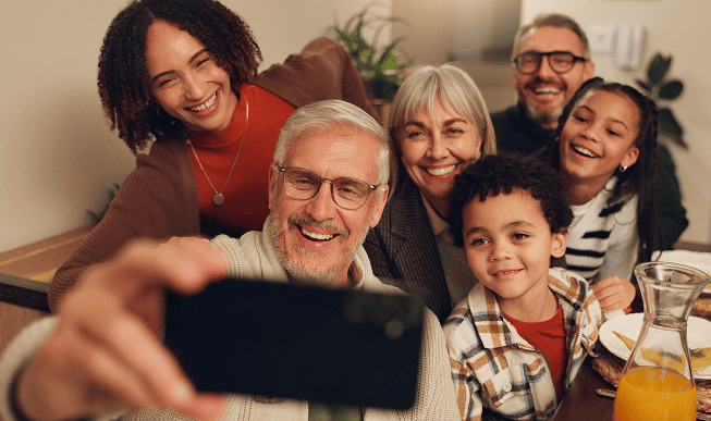 Smiling multigenerational family with caregivers and medicare beneficiaries taking a selfie together at a dining table.