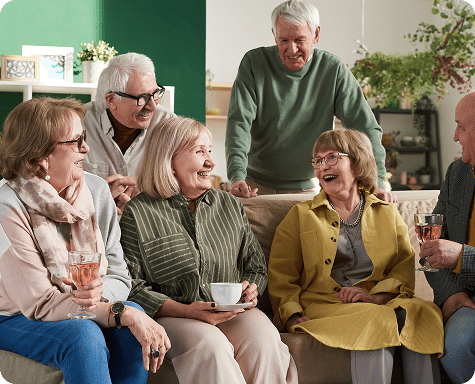 A group of six older adults enjoy discussion with primary care physician covered by medicare sitting together indoors, smiling and enjoying conversation with drinks.