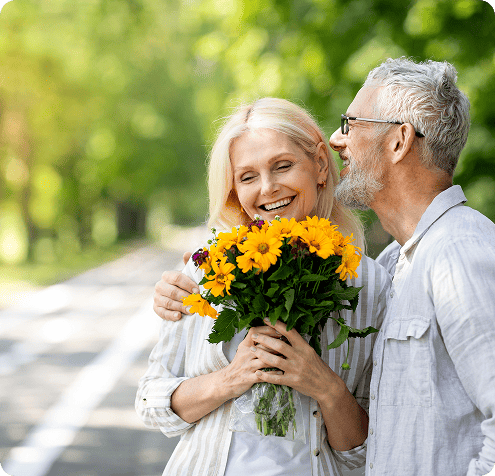 Smiling elderly couple on social security benefits misunderstand medicare advantage are outdoors with man presenting a bouquet of yellow flowers to the woman.
