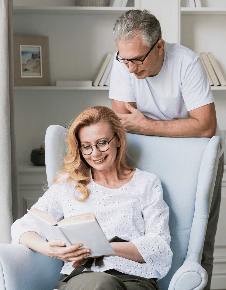 Smiling older woman with glasses reading a book in a light blue armchair while an older man stands behind her looking at the book.