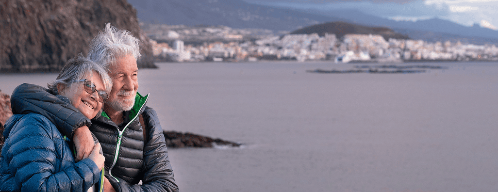 Smiling elderly couple in winter jackets hugging by a calm seaside with distant mountains and a town in the background.