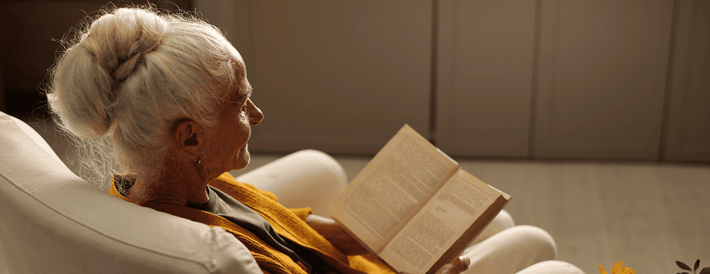 Elderly woman with white hair in a bun, sitting on a beige chair, reading a book indoors.