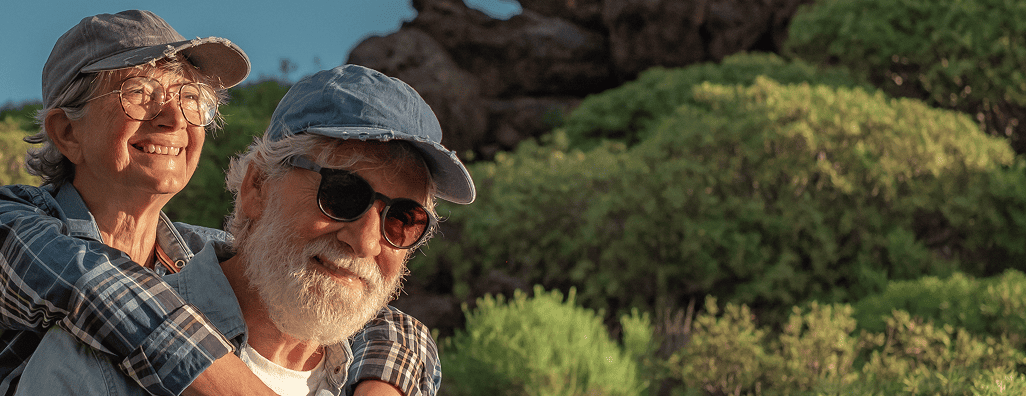 Smiling elderly couple outdoors with lush green plants in the background, both wearing caps and glasses.