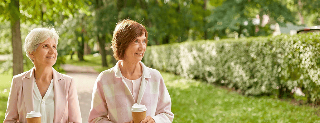 Two middle-aged women walking outdoors in a park, each holding a coffee cup and smiling.