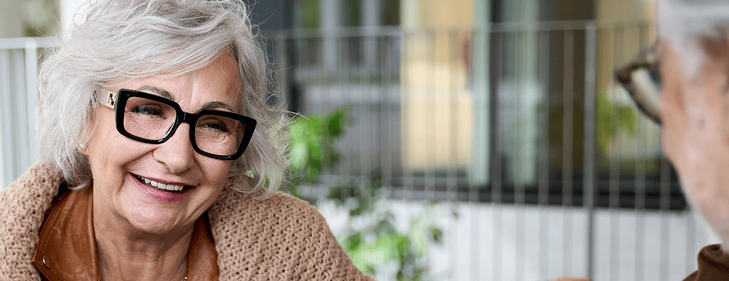 Smiling elderly woman with white hair and large black glasses conversing with another person.