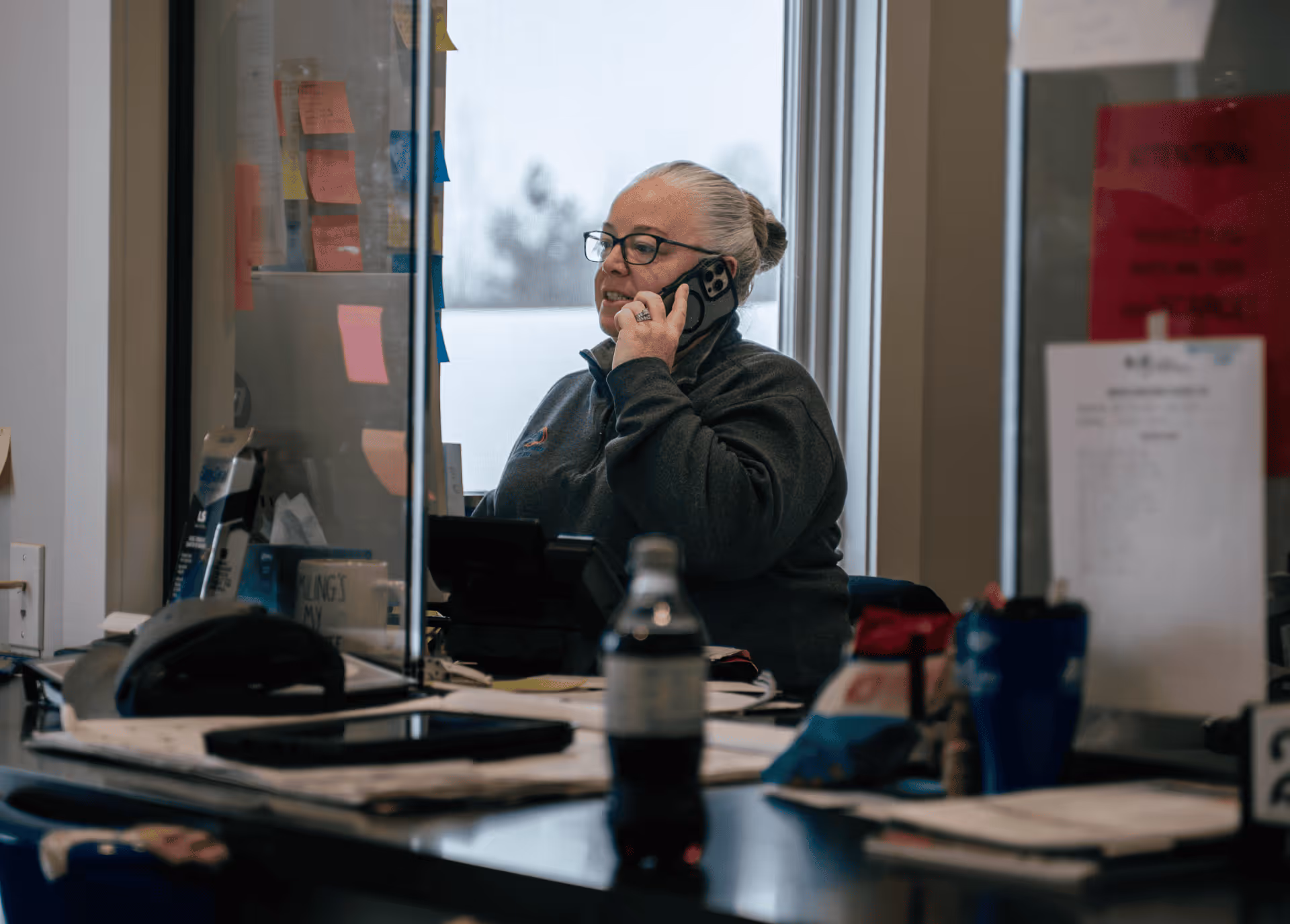 Woman with glasses and gray hair tied back talking on a smartphone in an office with sticky notes on a glass partition.