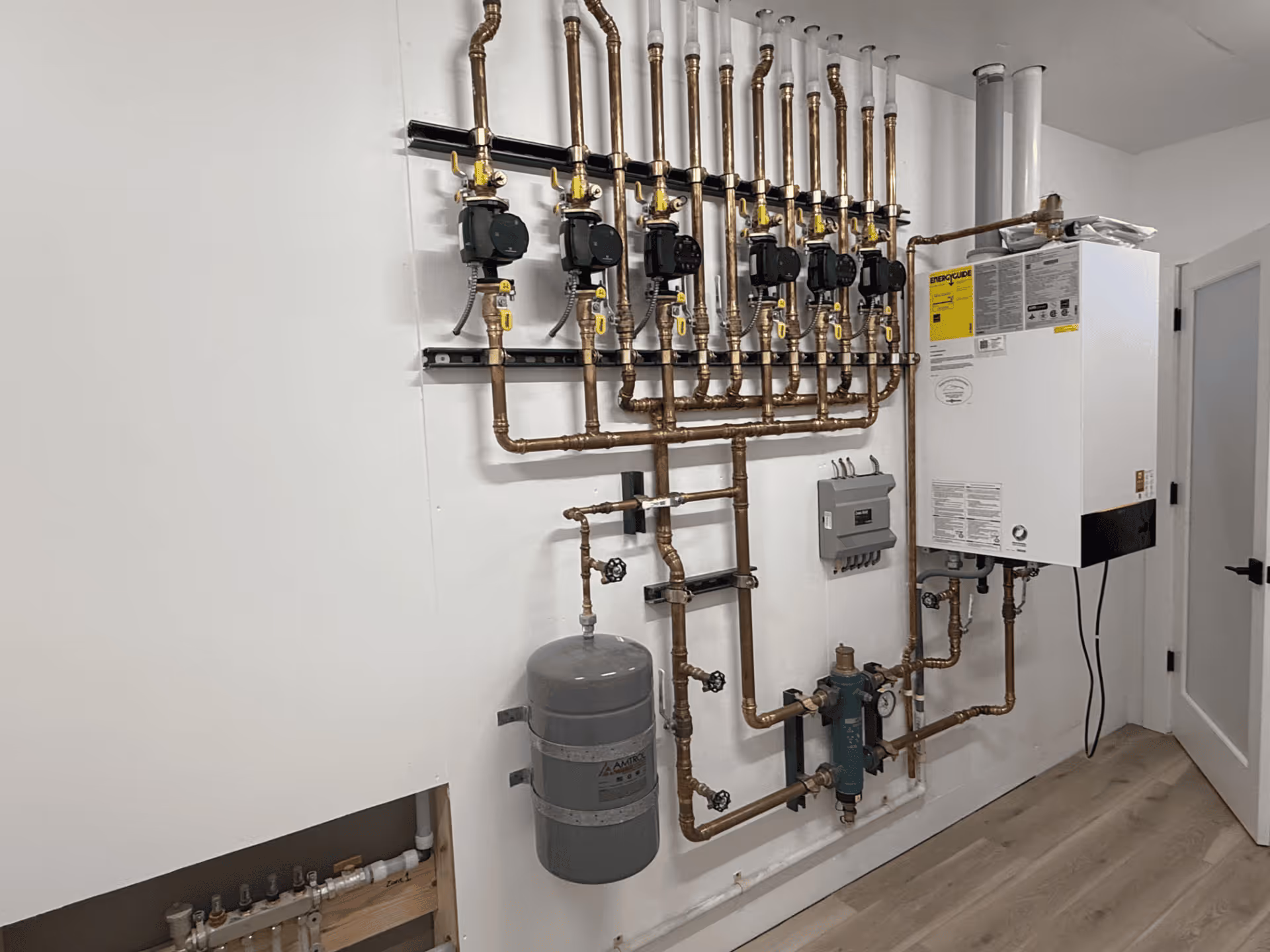 Complex plumbing and heating system with multiple copper pipes, valves, a gray expansion tank, and a white wall-mounted boiler unit in a utility room.