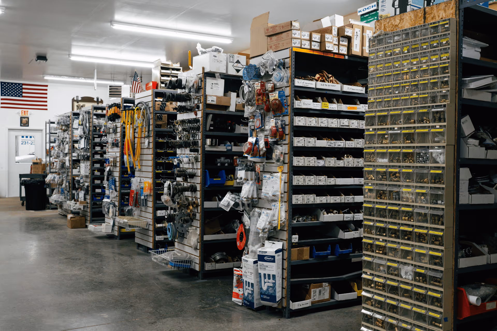 Interior of a hardware store with shelves stocked with various tools, parts, and boxes, and an American flag on the back wall.
