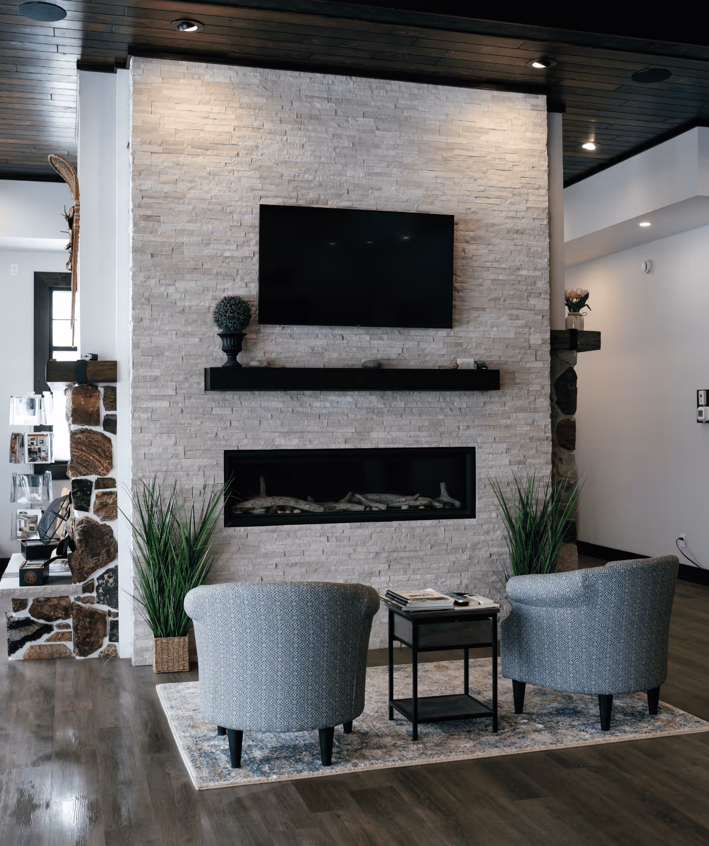 Modern living room with two patterned armchairs, a small black side table, a wall-mounted flat screen TV, and a linear gas fireplace set in a white stone accent wall.