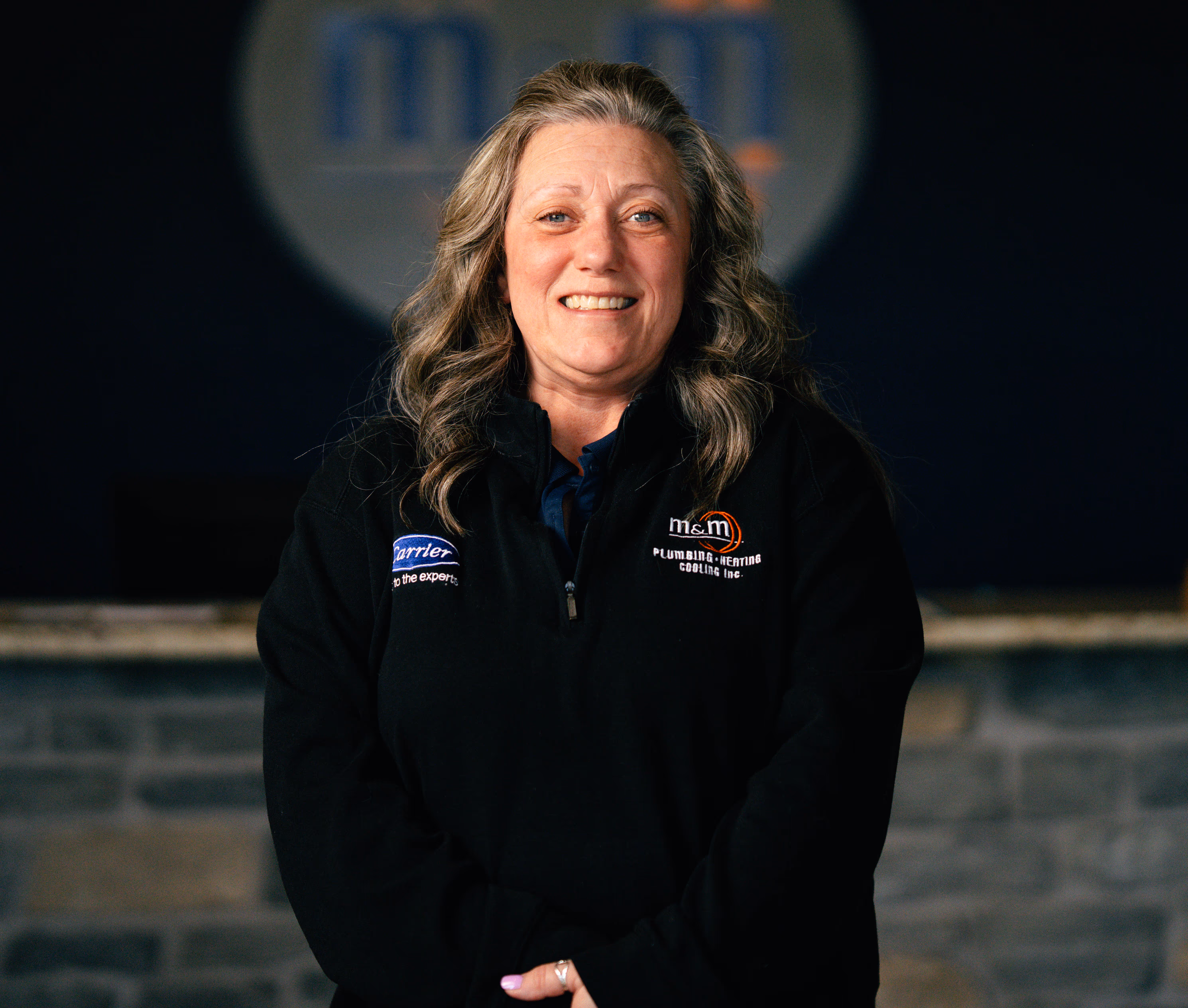 Smiling woman with gray wavy hair wearing a black M&M Plumbing Heating Cooling jacket in a workplace setting.