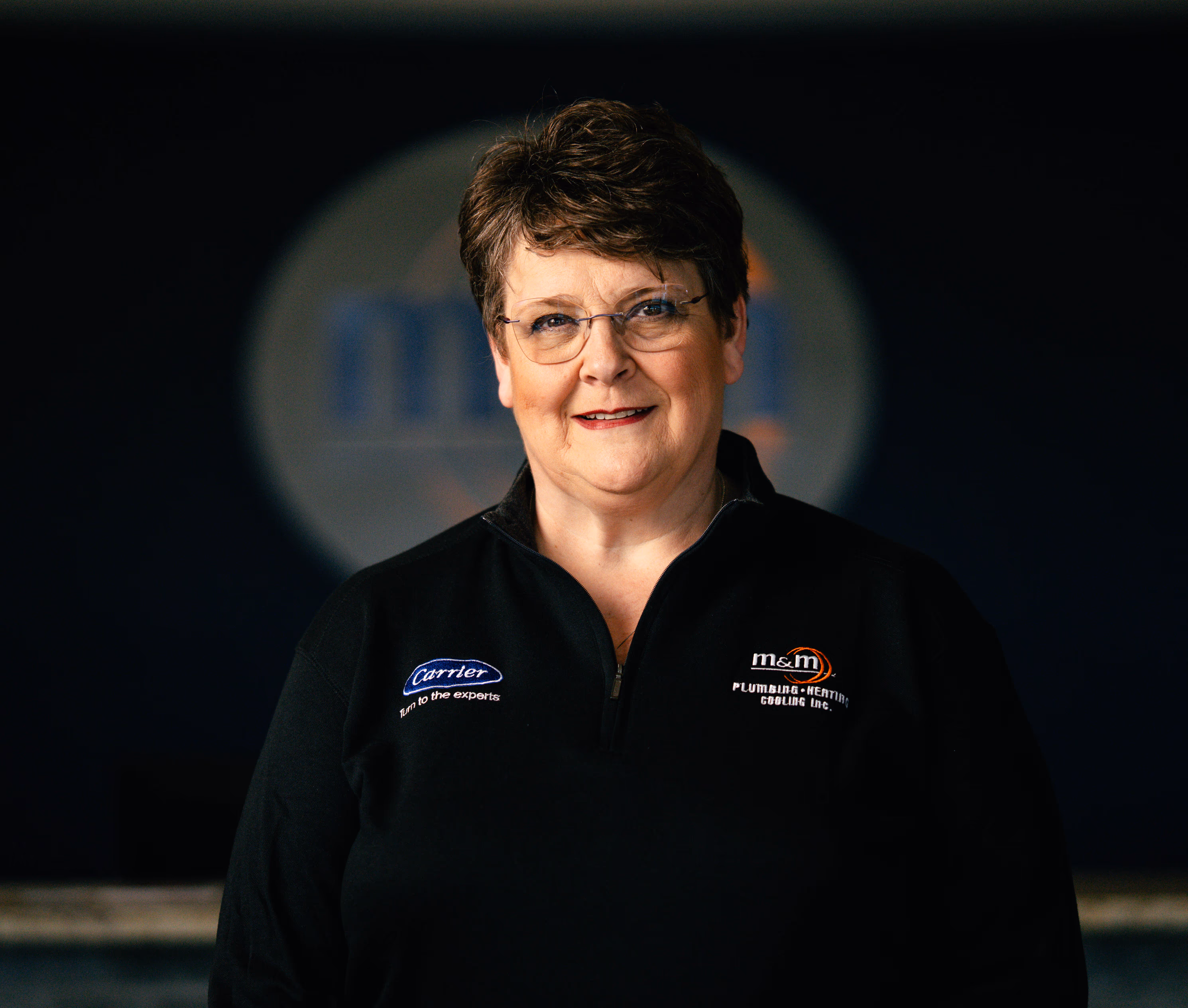 Smiling woman with short hair wearing glasses and a black jacket with Carrier and m&m Plumbing logos.