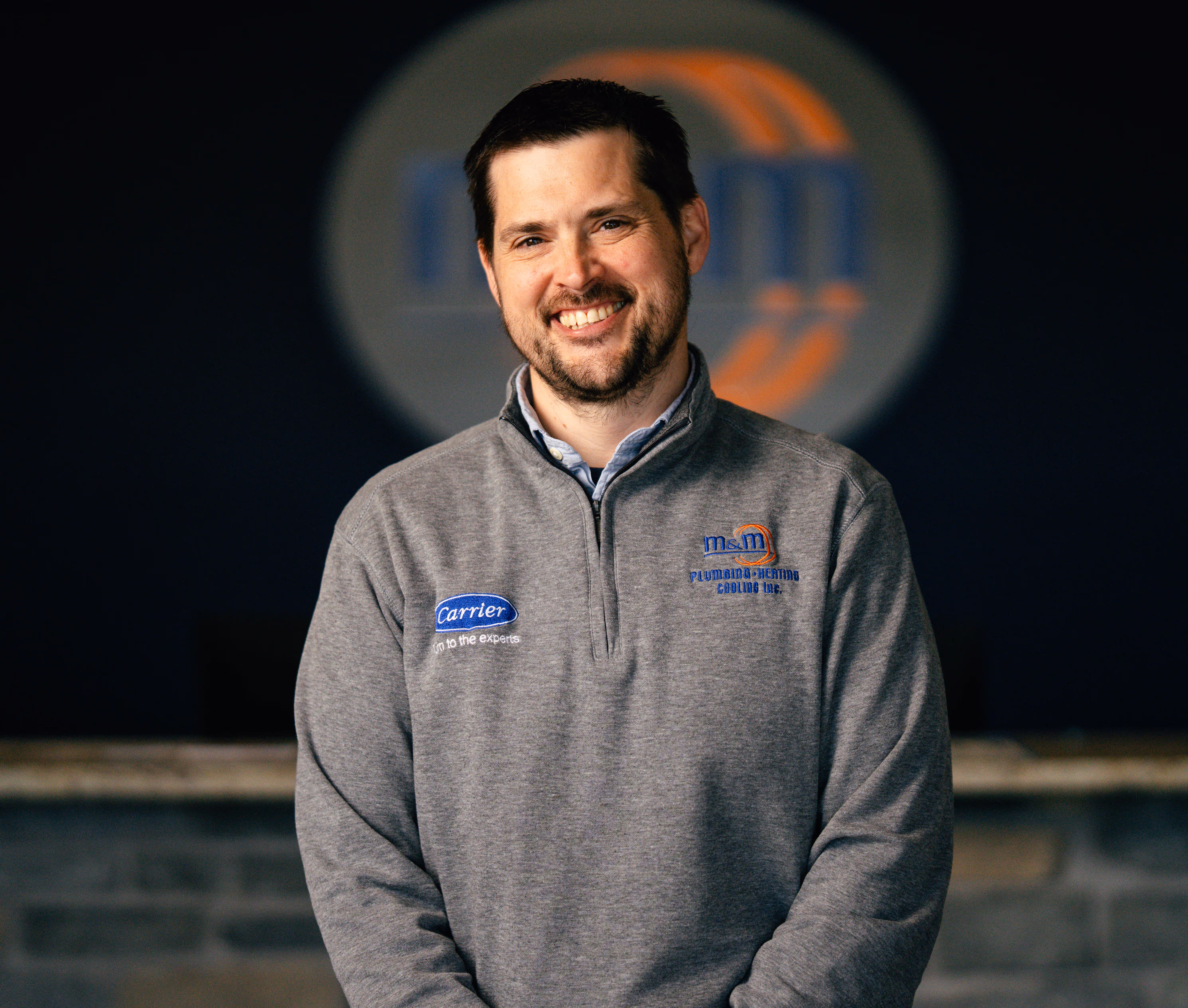 Smiling man wearing a gray pullover with plumbing and heating company logos standing in front of a dark background.