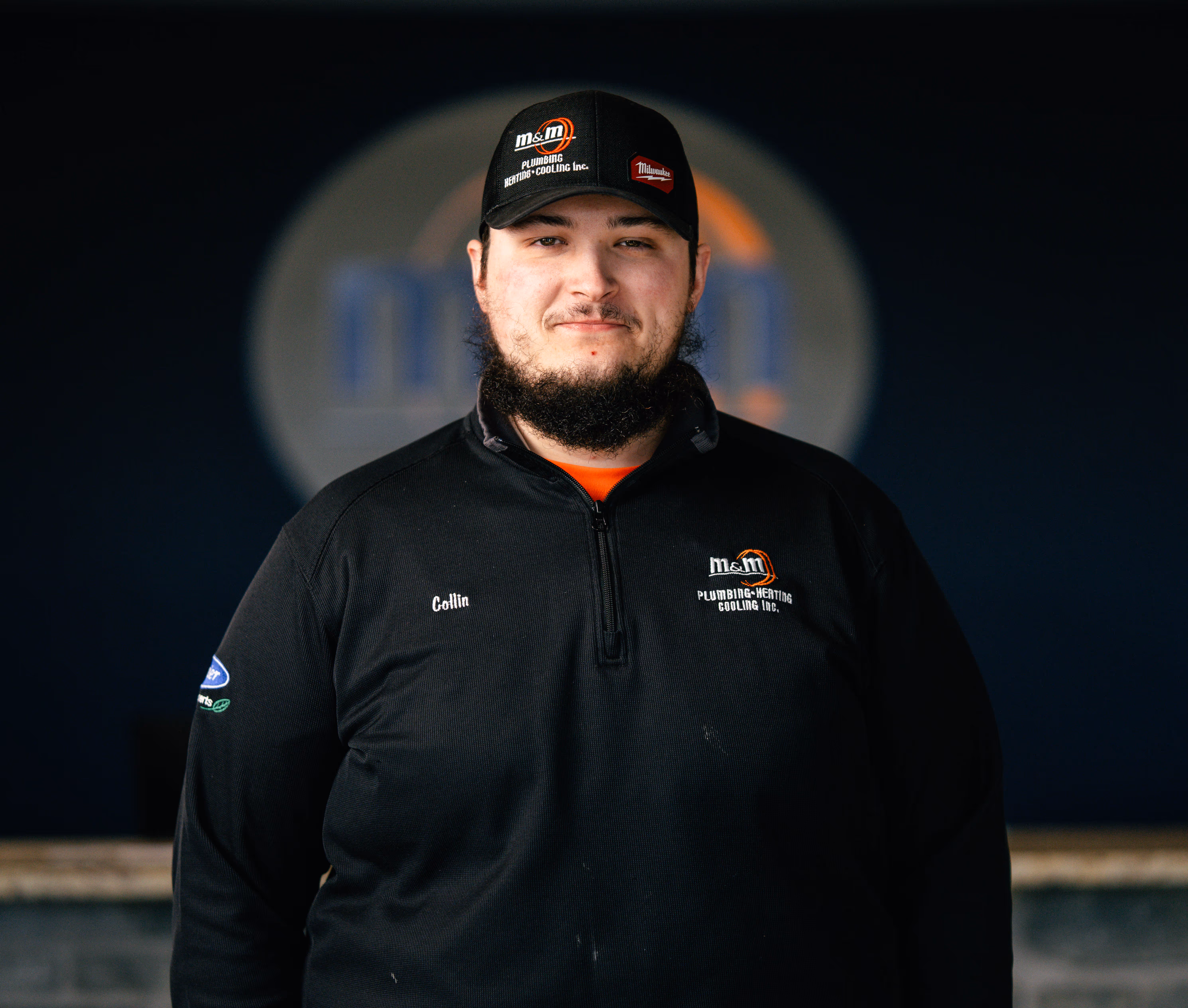Man wearing a black M&M Plumbing Heating Cooling Inc. jacket and cap standing indoors.