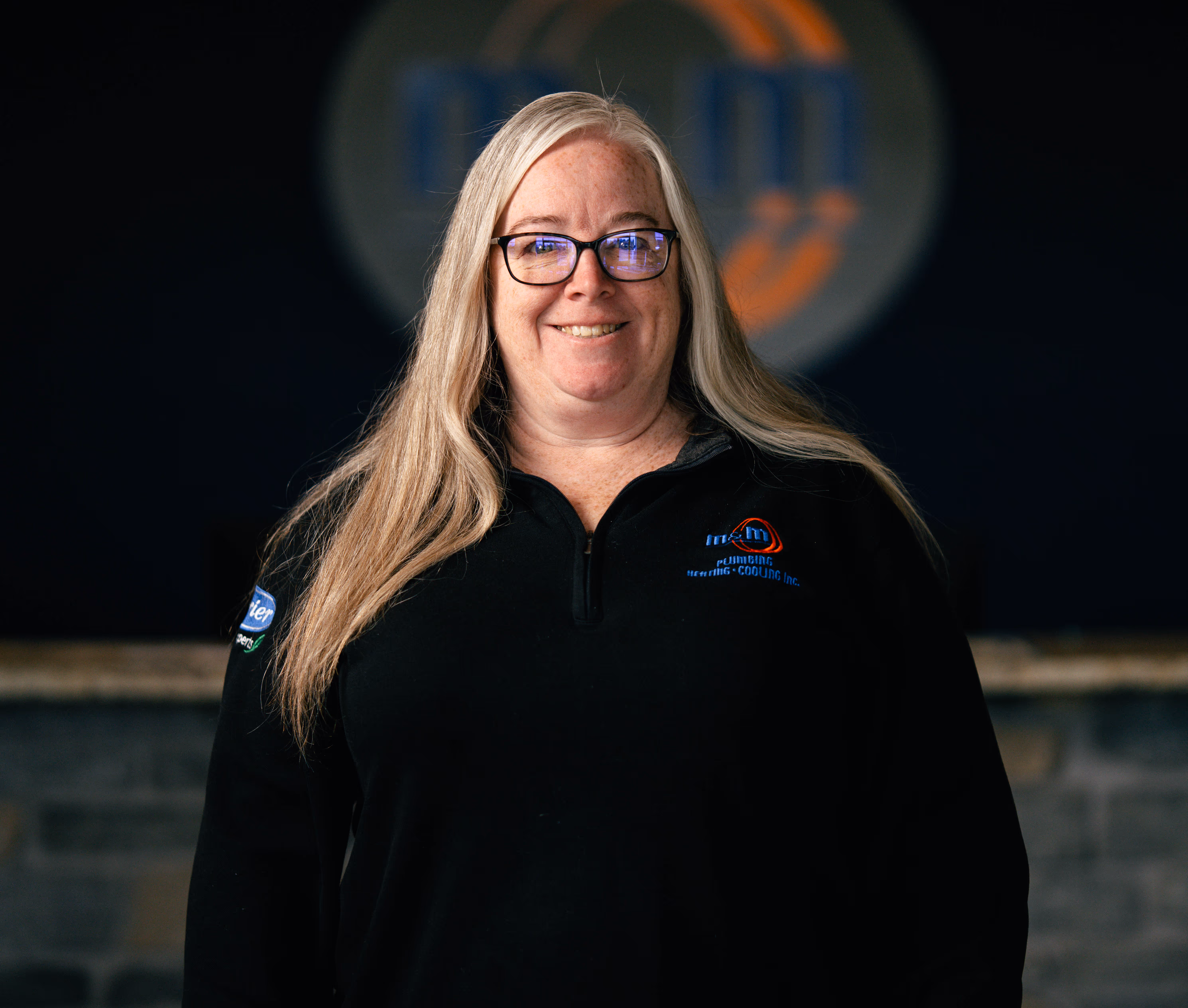 Woman with long blonde hair and glasses smiling, wearing a black quarter-zip shirt with a company logo.