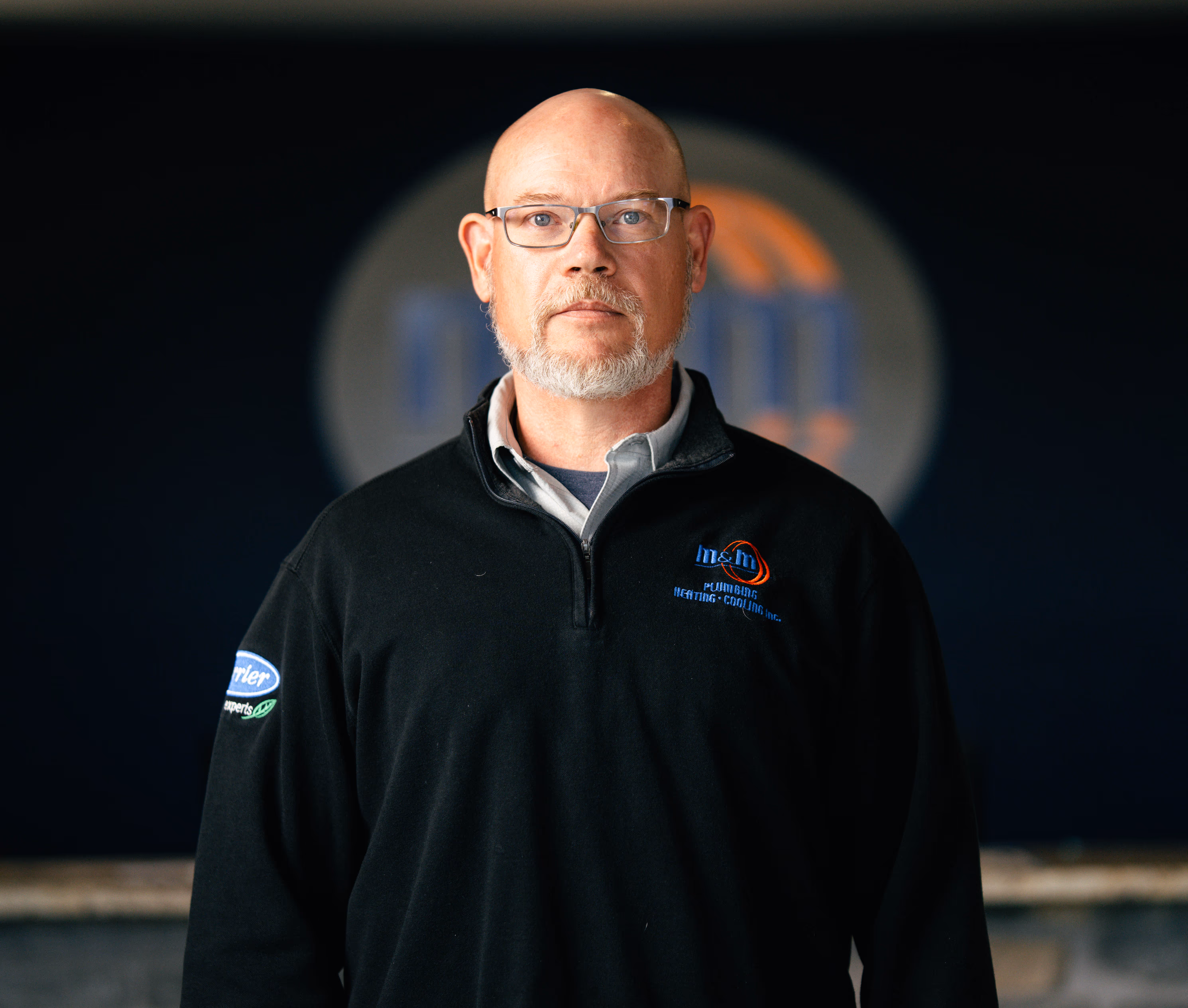 man with glasses and a beard wearing a black M&M Plumbing Heating Cooling pullover.