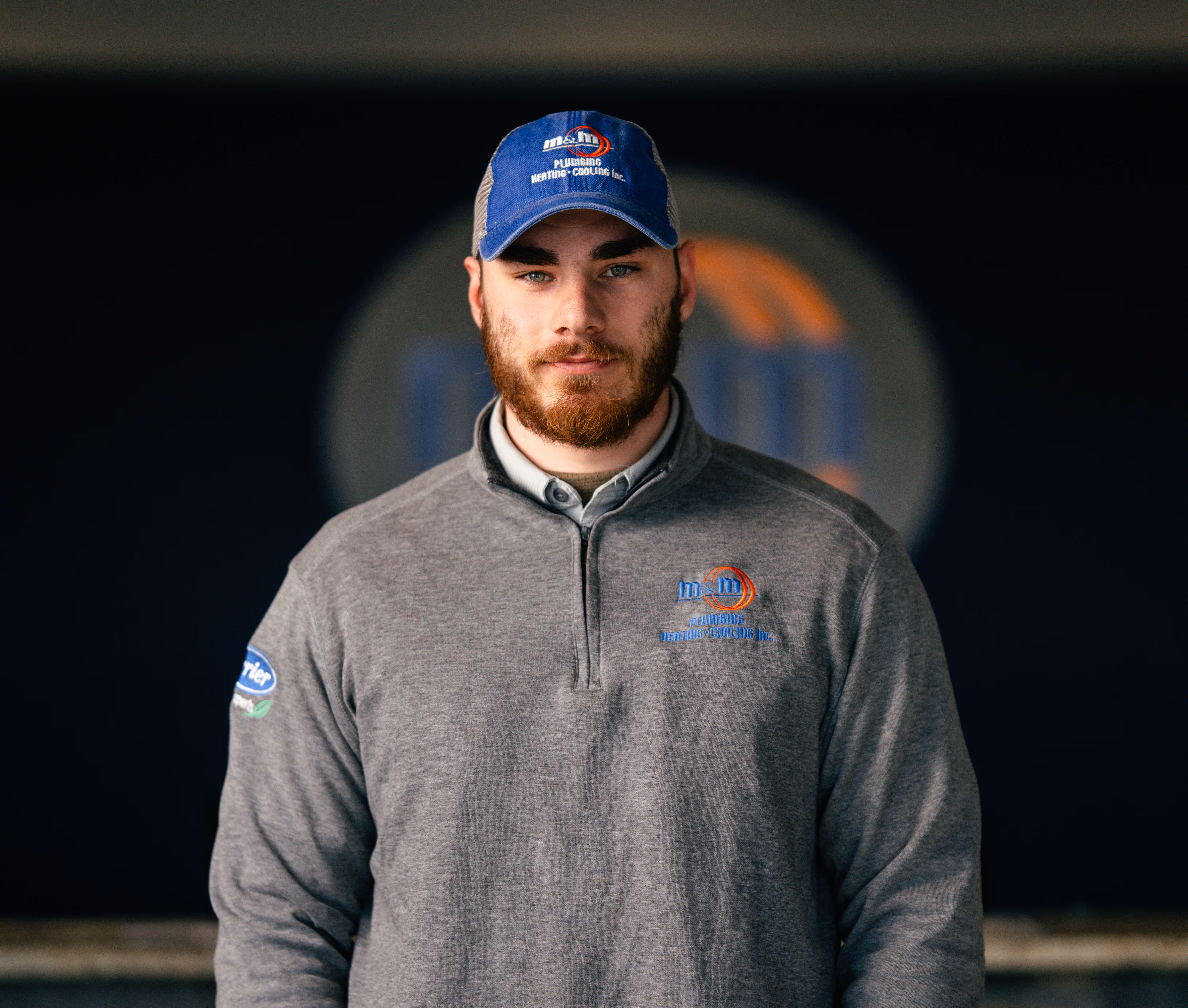 Bearded man wearing a blue cap and gray pullover with M&J Plumbing Heating Cooling Inc. logo.