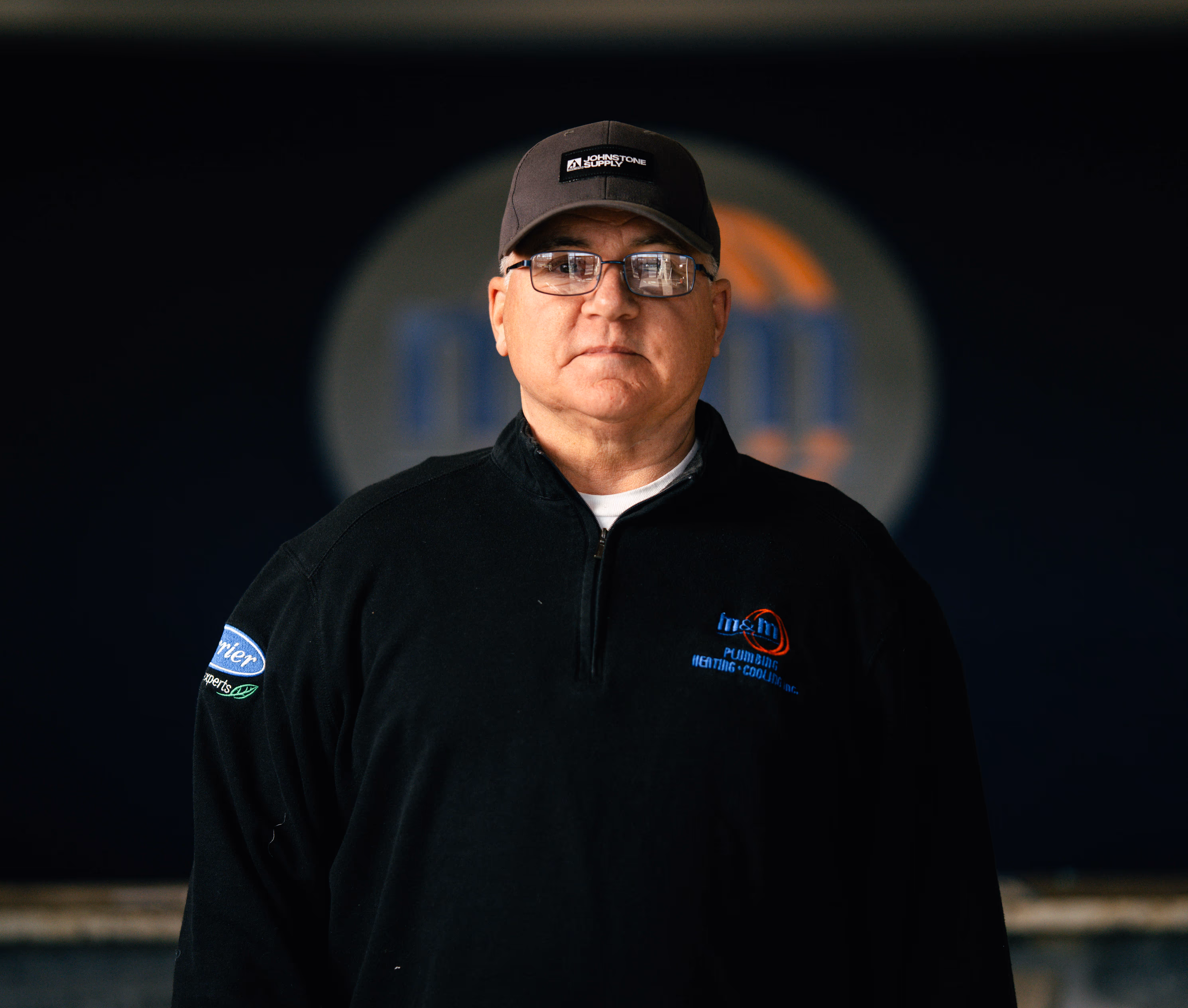 Man wearing glasses, a black Johnstone Supply cap, and a black sweater with an m&m Plumbing Heating Cooling logo on the chest.