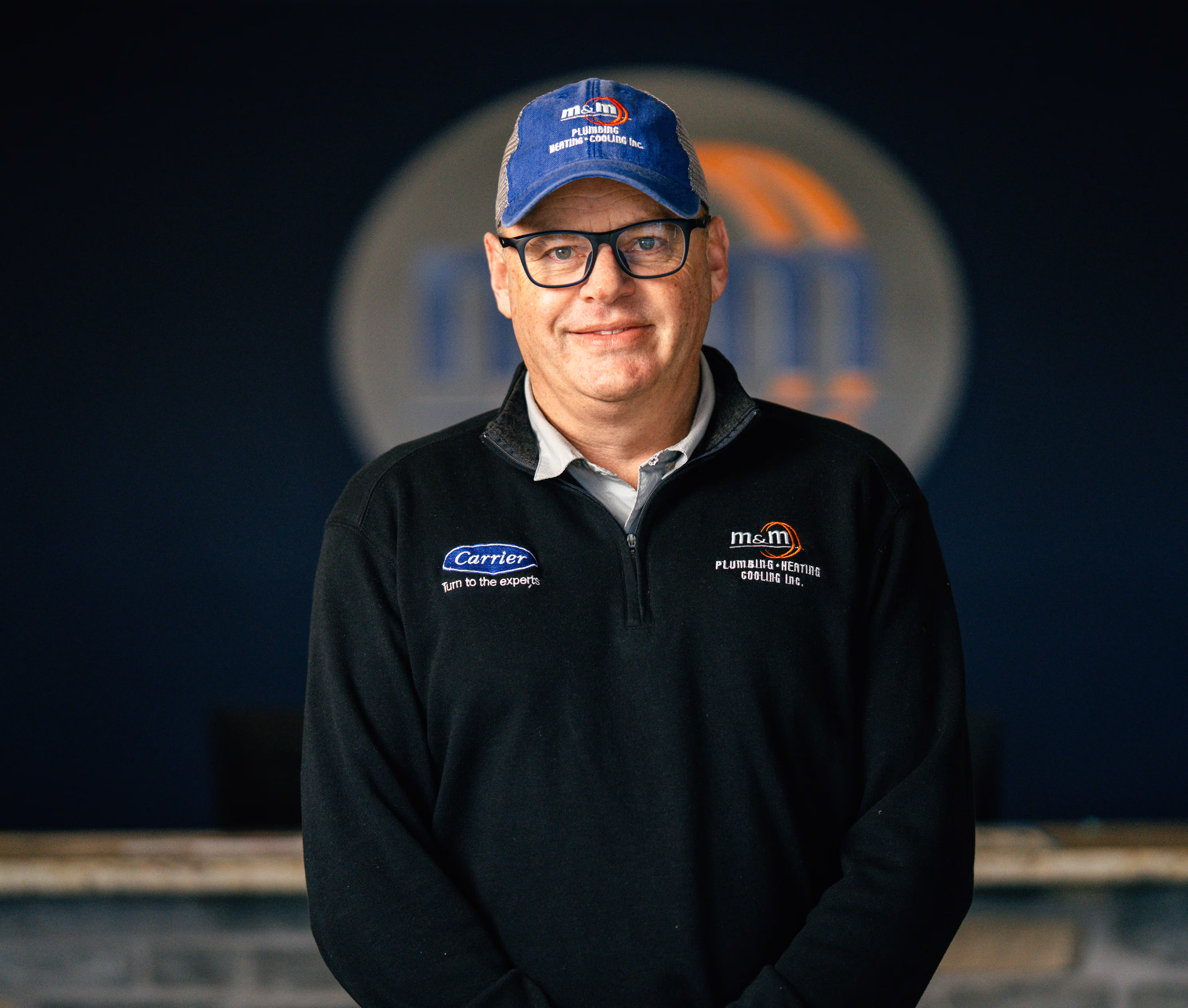 Man wearing glasses, blue M&M Plumbing Heating Cooling Inc. cap, and black jacket with company logos standing in front of a blurred circular logo.