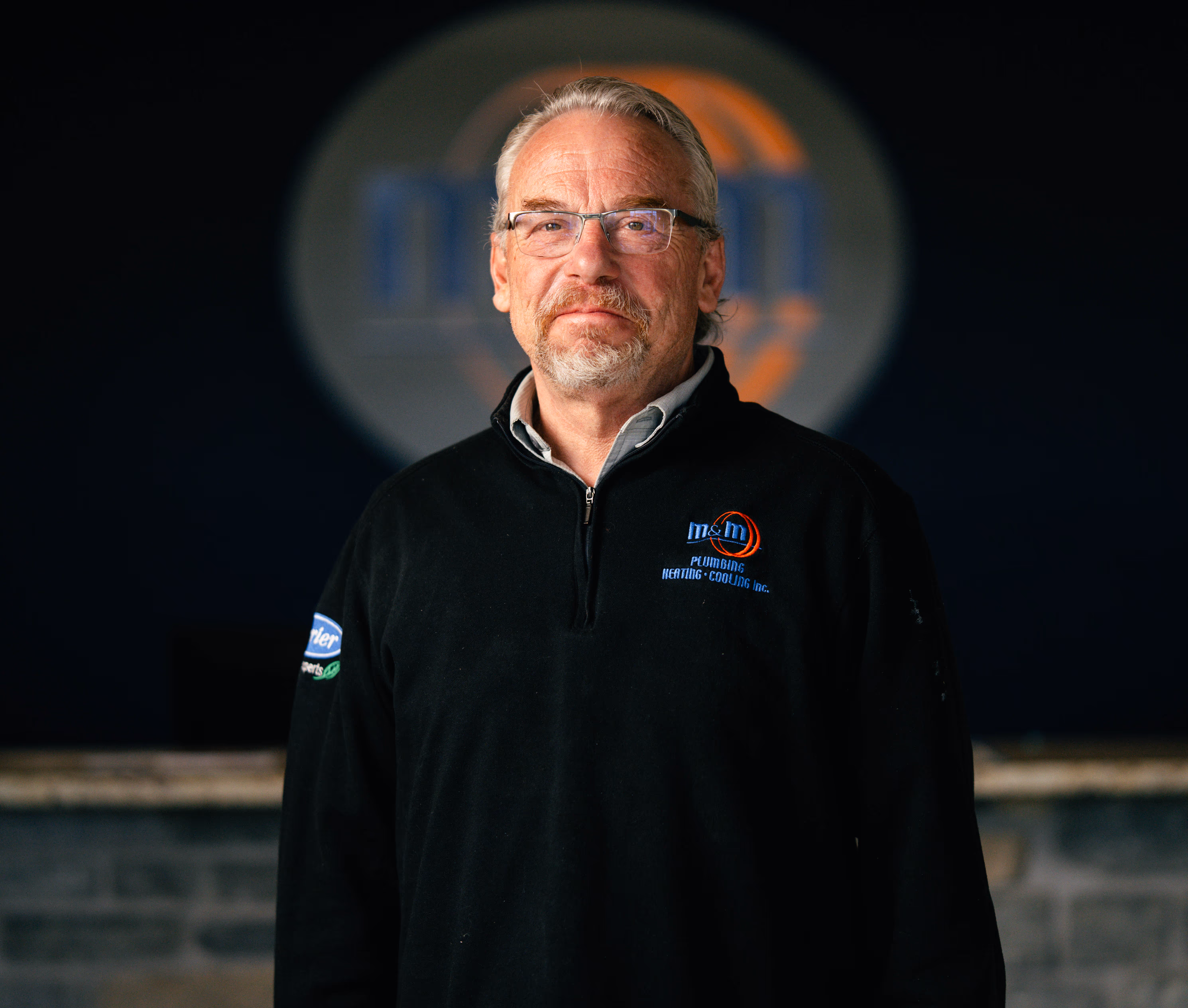 man with glasses and gray hair wearing a black zip-up with a plumbing, heating, and cooling company logo.