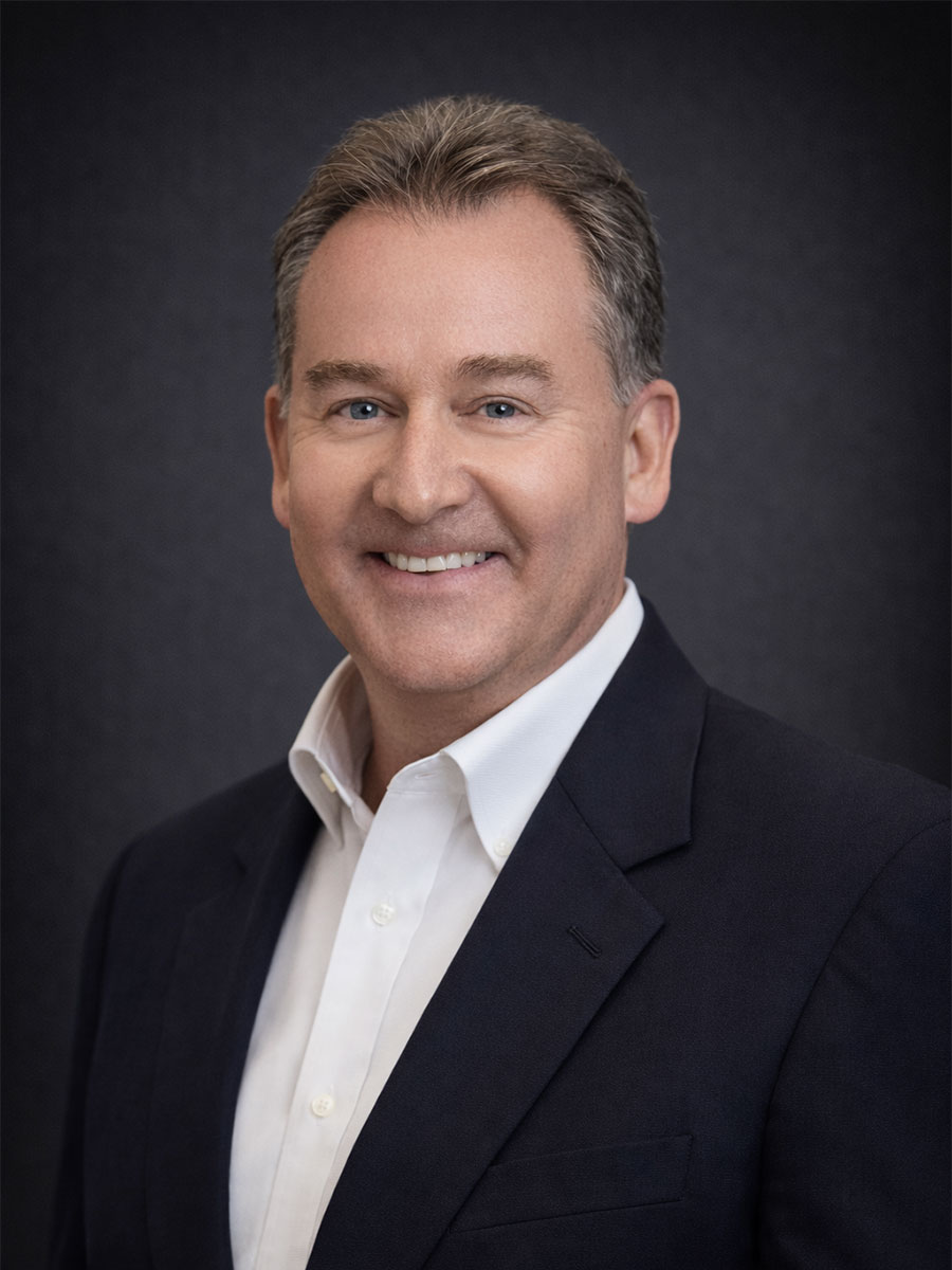 A middle-aged man in a dark suit and white shirt smiles in front of a plain dark background.
