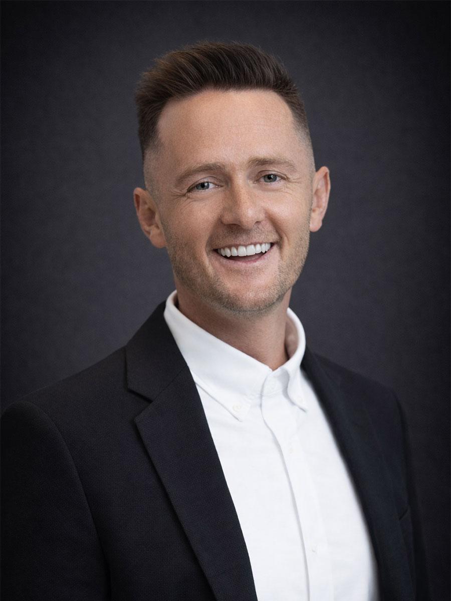 Smiling man in a black blazer and white shirt poses against a dark, plain background.