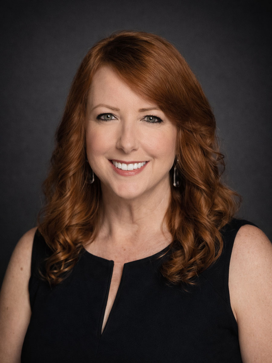 Woman with long red hair and a sleeveless black top smiling in front of a dark background.